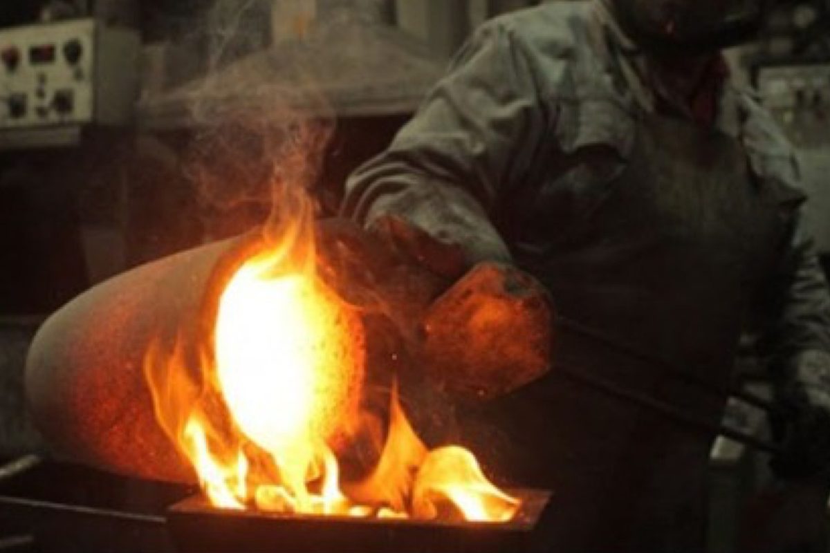 Operador vazando ouro fundido de um cadinho para a forma, com chama alta, dentro da refinaria